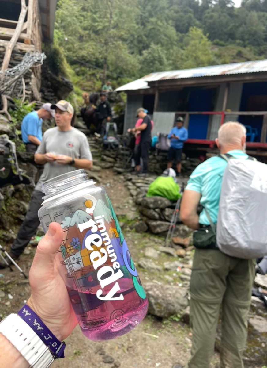 A trekker holds up an 'immune ready' water bottle on a mountain mission trip, with team members and a trail lodge behind.