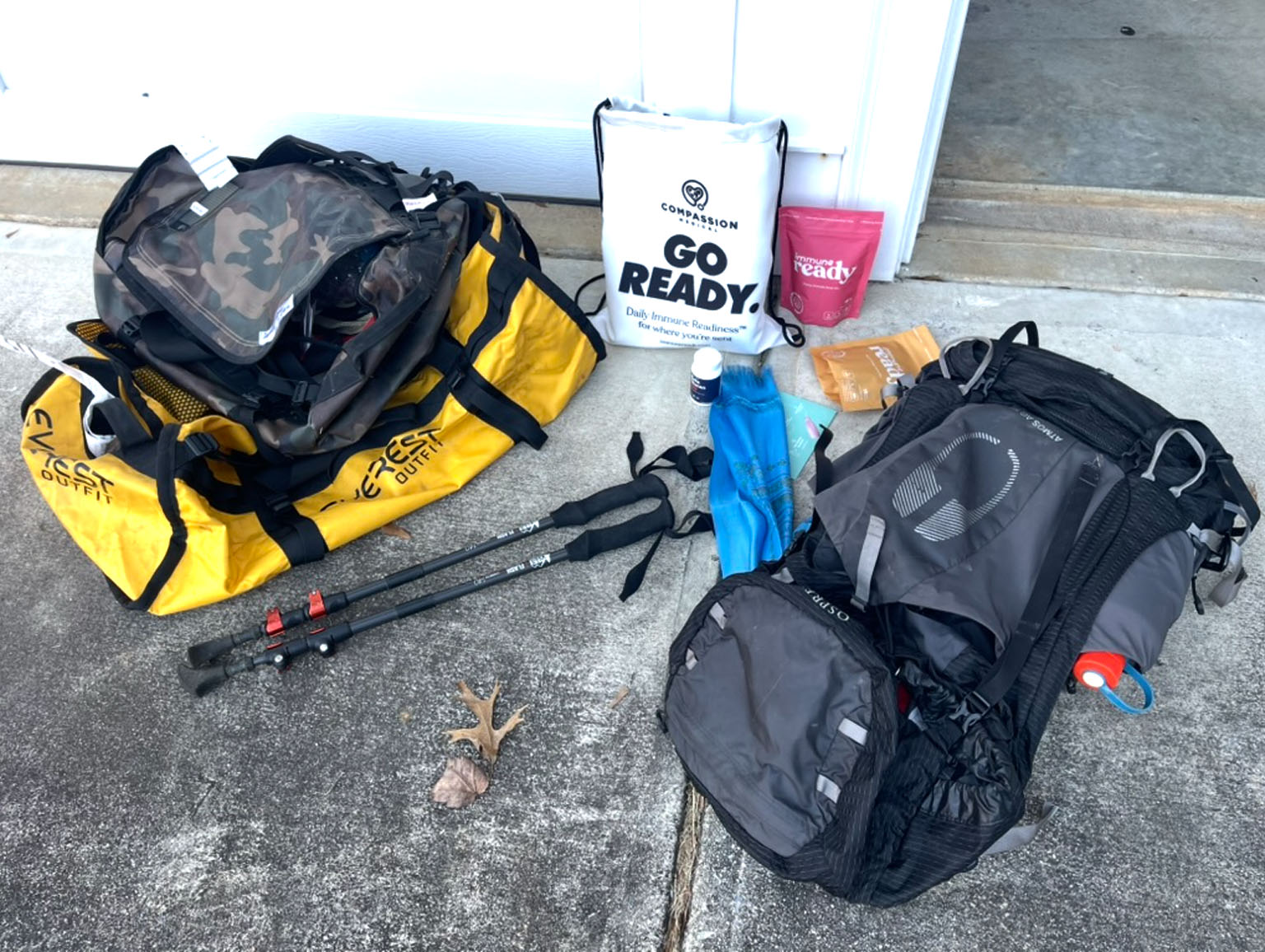 An expedition go-bag flat-lay — a duffel, backpack, and trekking poles beside an Immune Ready 'Go Ready' daily immune-readiness pack and pouches.