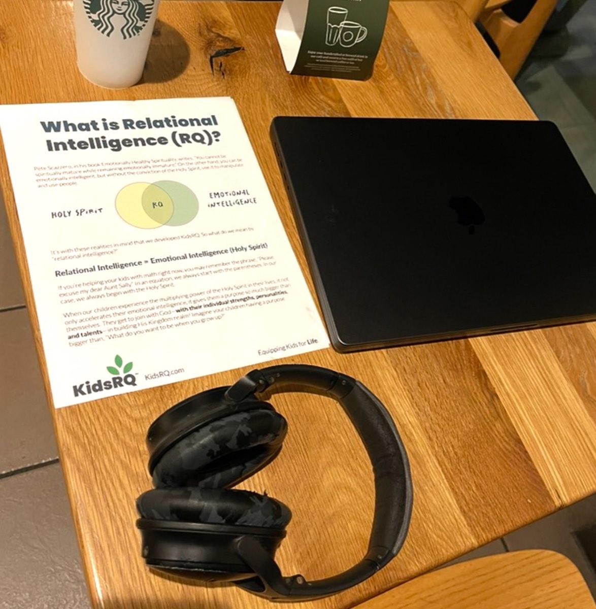A KidsRQ 'What is Relational Intelligence?' handout with its RQ Venn diagram on a café worktable, beside a laptop and headphones.