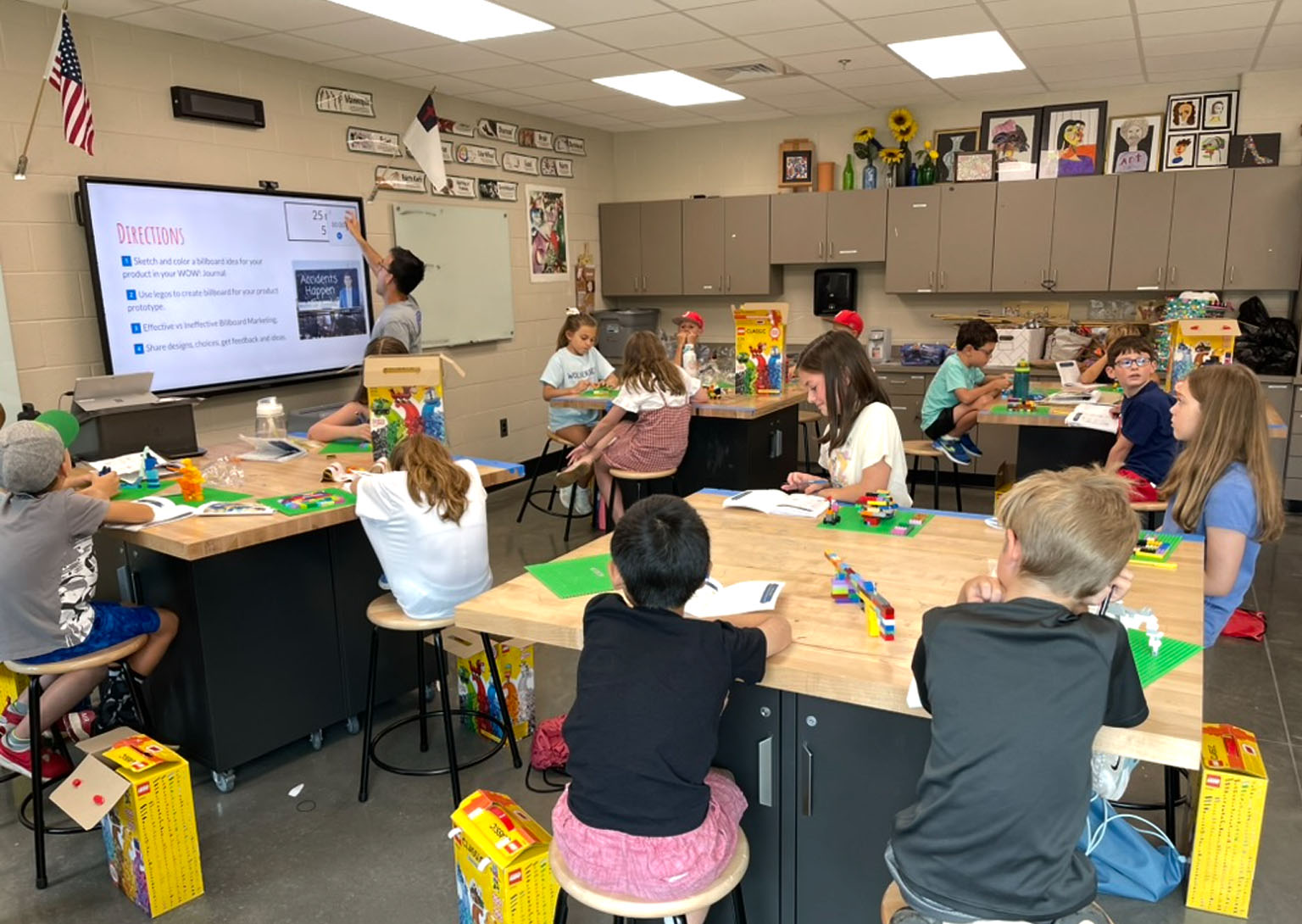 A WOW Camp maker-space classroom — kids at worktables building product prototypes with LEGO while an instructor leads at the screen.